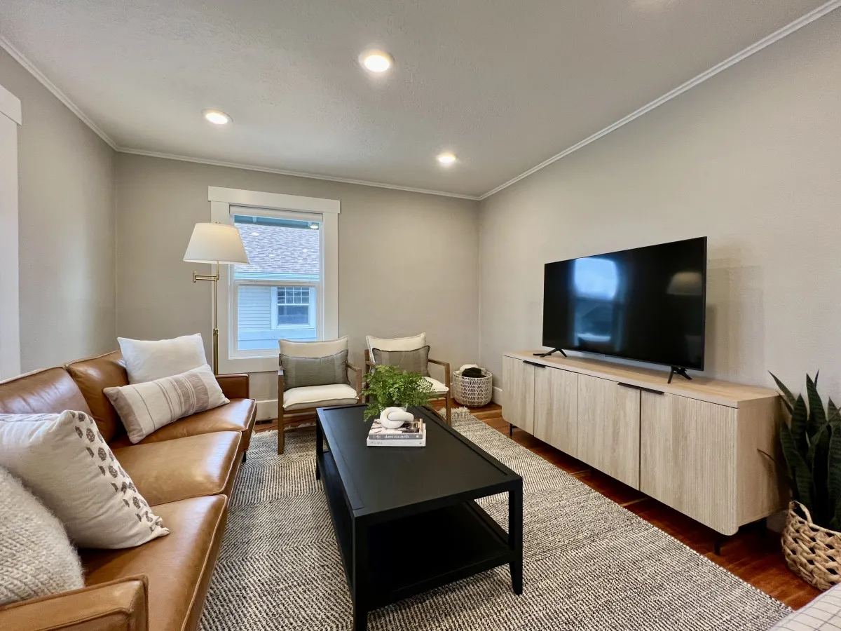 A cozy interior featuring a leather sofa, black coffee table, television, and decorative plants. The space is bathed in gentle sunlight, enhancing its welcoming vibe for relaxing, reading, or spending time with loved ones—including furry companions.