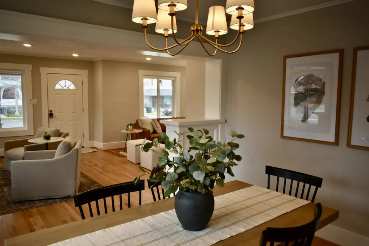 A wide, inviting view of the main living area featuring a chandelier overhead, a dining table adorned with green plants, and a plush, comfortable seating area. The space exudes warmth and is designed for both intimate dinners and relaxed gatherings, with original architectural charm evident in the fixtures and layout.