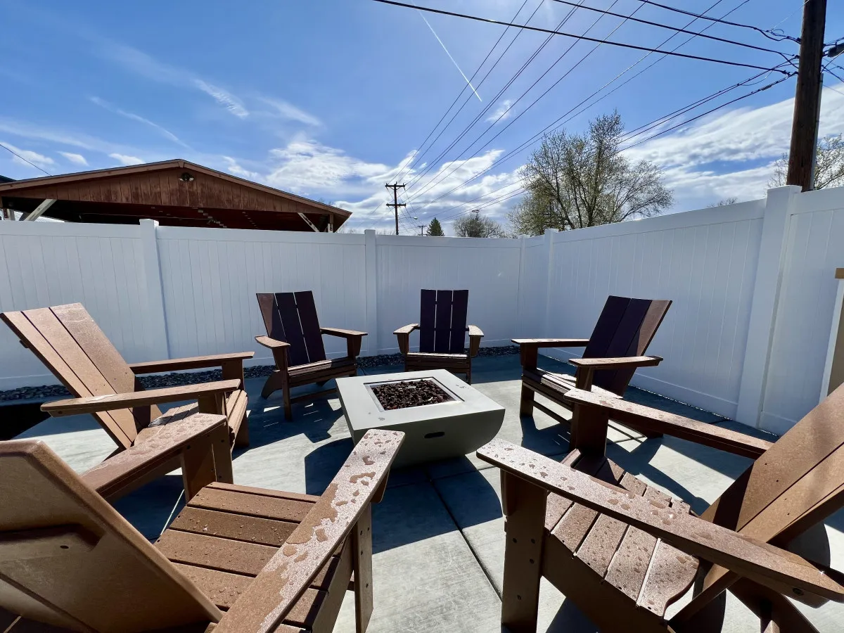A cozy exterior scene featuring a fire pit surrounded by Adirondack chairs and enclosed by a white fence. The space is designed for evening gatherings, socializing, and relaxation under the stars, creating a welcoming atmosphere ideal for large and small groups.