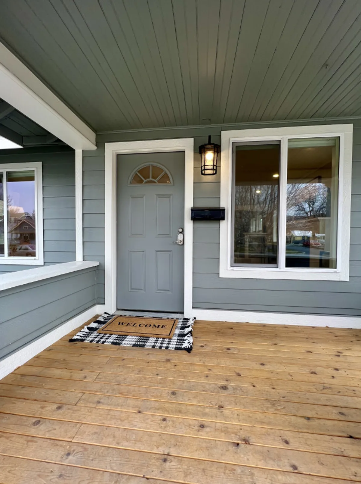 The image shows the inviting gray front door of The Winston, accented by a welcome mat, lantern-style light fixture, and large windows. The porch area is visible, suggesting a warm welcome for guests and pets alike.
