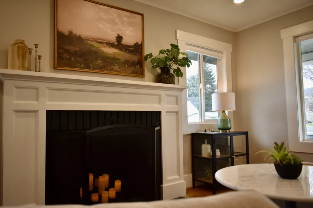A welcoming living room with a glowing fireplace, a landscape painting above the mantle, a lush potted plant, a table lamp casting a soft glow, and a glass-front cabinet. The space feels intimate and perfect for unwinding after an adventurous day.