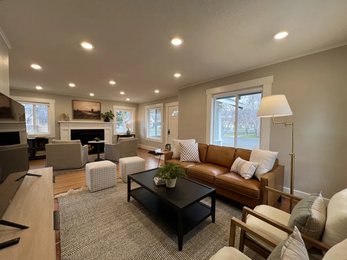 A cozy living area featuring a brown leather sofa, black coffee table, large windows streaming in sunlight, and artwork above a classic fireplace. The setting is both heritage-rich and updated for comfort, ideal for relaxing or entertaining guests.