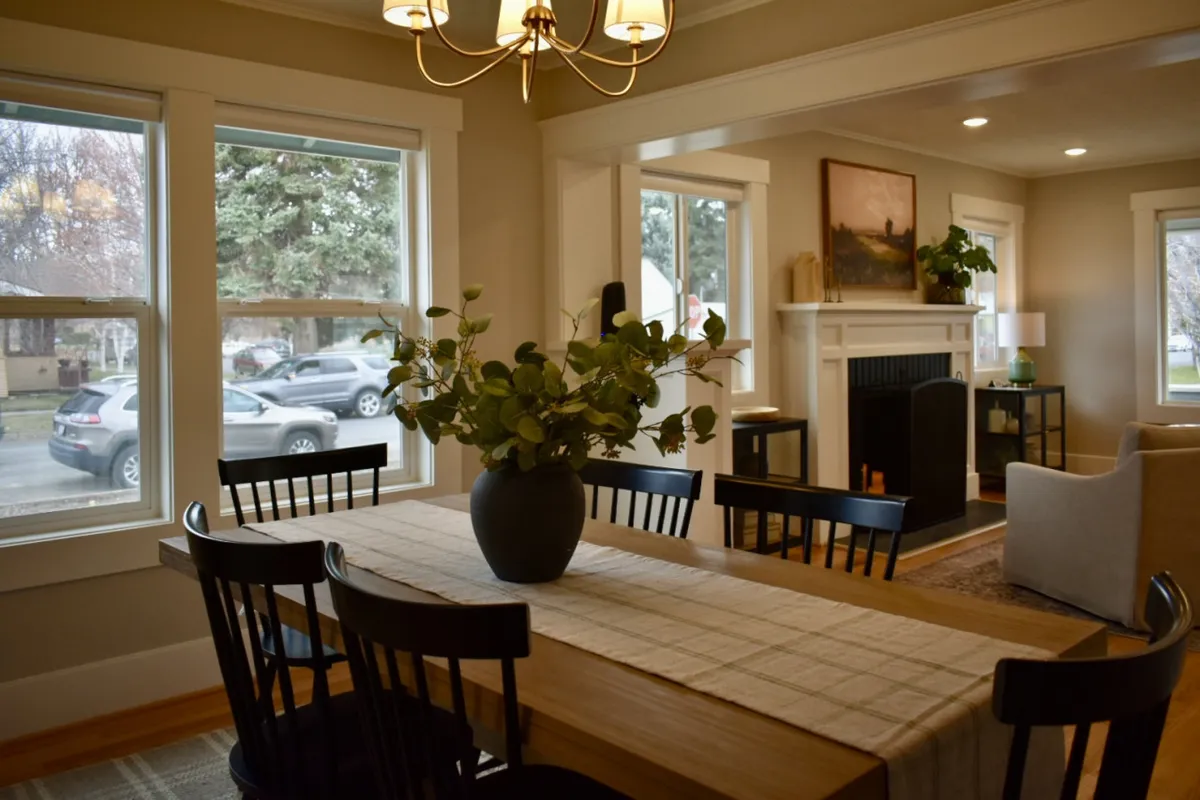A bright, inviting dining area featuring a large table with a vase of greenery, expansive windows filling the space with natural light, a classic fireplace, and a comfortable seating area. The room feels ideal for shared meals or planning the next day's adventure.